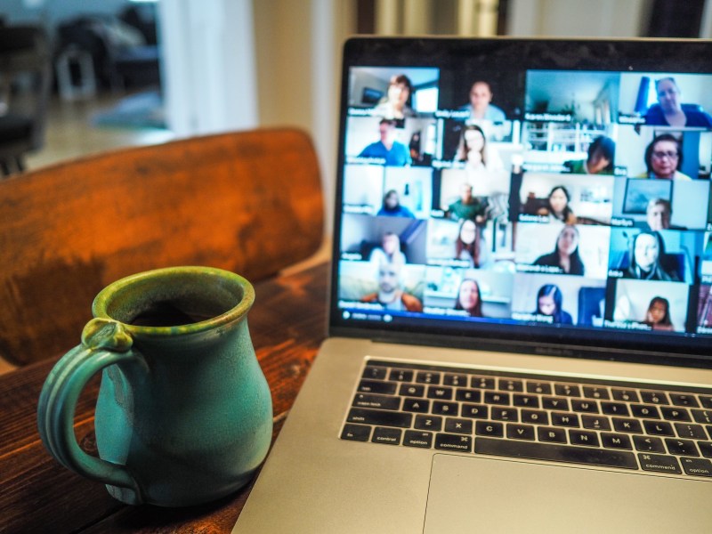 A laptop screen displaying a group of people on a zoom call, on a table next to a mug.
