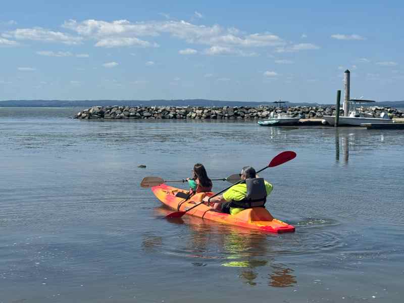 Orange kayak in a calm part of the Chesapeake Bay. There are two kayakers. Nick sits in the back with a neon green shirt and blue life vest. A woman sits in the front with a red life vest and paddle held horizontally in an adaptive clamp mounted to the bottom if the kayak seat.