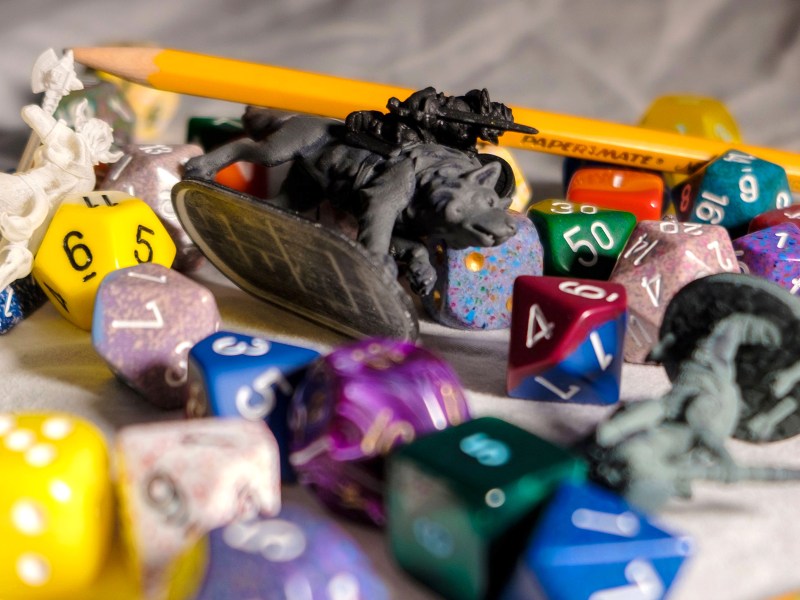 A variety of tabletop gaming pieces scattered on a table, including different types of dice in different colors, a figurine of a warrior riding a wolf, a yellow pencil, and blurry figurines of characters carrying weapons